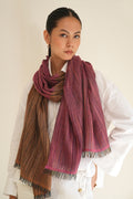 A woman wearing a white button-up shirt and white pants, styled with a long fringed scarf in a dusty rose and brown striped pattern, against a plain cream background.