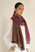 A woman wearing a white shirt and a large woven scarf with fringed edges, draped around her neck. The scarf has a plaid pattern in brown, burgundy, and muted pink tones, photographed against a plain beige background.