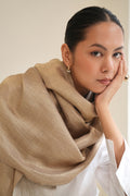 A woman with dark hair pulled back wears a soft gold metallic and beige shawl draped over her shoulders and a white collared shirt. She is posed in profile with her hand resting on her cheek, wearing small gold hoop earrings and a gold ring, against a plain light beige background.