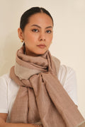 A woman wearing a rose gold and natural cashmere wrapped around her neck, paired with a white t-shirt and small gold earrings, set against a plain beige background.