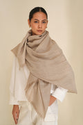A woman modeling a large, metallic silver and beige cashmere shawl with fringed edges, draped elegantly over her shoulders and wrapped around her body, paired with a white top and white trousers, against a solid light beige background.
