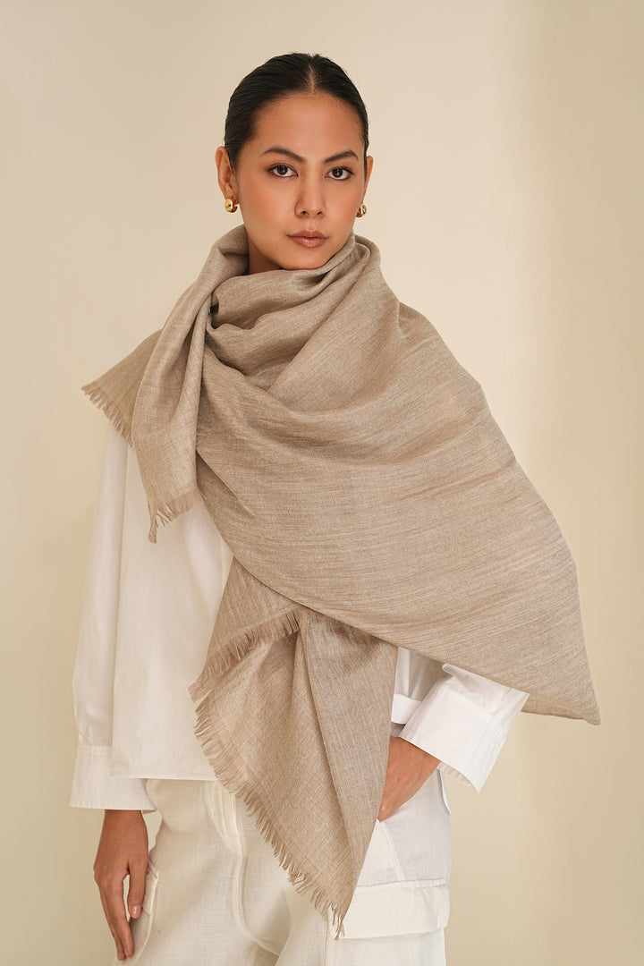 A woman modeling a large, metallic silver and beige cashmere shawl with fringed edges, draped elegantly over her shoulders and wrapped around her body, paired with a white top and white trousers, against a solid light beige background.