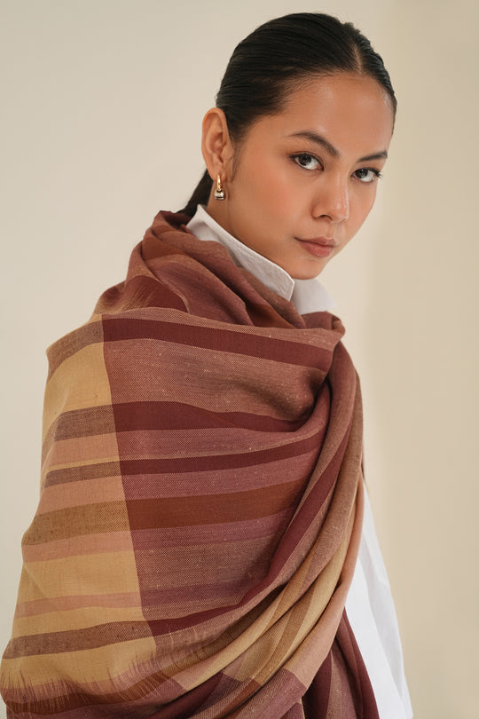 A woman wearing a striped shawl in earthy tones of brown, rust, and beige over a white collared shirt, photographed against a plain light beige background.
