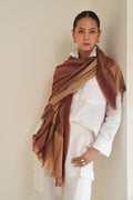 A woman wearing a white outfit and a large striped shawl scarf in shades of brown, rust, and beige, standing against a plain light beige wall.
