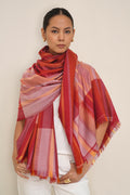 A woman modeling a large fringed shawl with a plaid pattern in red, orange, pink, and purple tones, styled over a white top and white pants against a plain beige background.