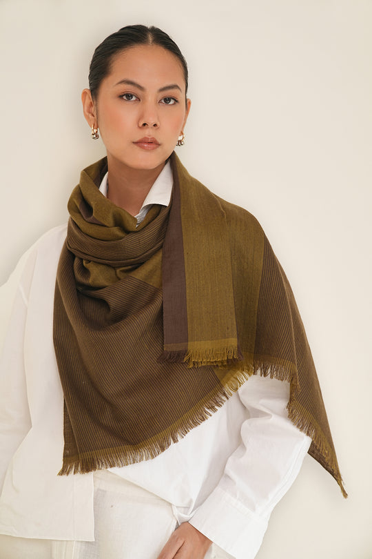 A woman wearing a large olive-brown fringed shawl draped over her shoulders, styled over a white collared shirt against a plain off-white background.