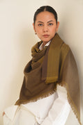 A woman wearing a large olive-brown and brown fringed shawl over a white top, posed against a plain off-white wall.